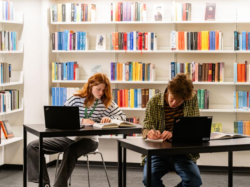 To unge sidder ved hvert deres bord og arbejder med bøger og computere. De sidder på et bibliotek. 